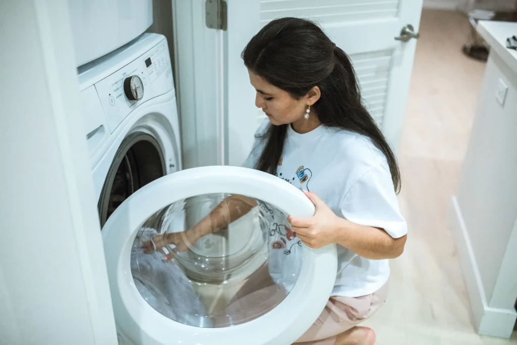 Woman kneeling beside front-load washing machine, opening the door to load laundry — trusted clothes washing service in Doha