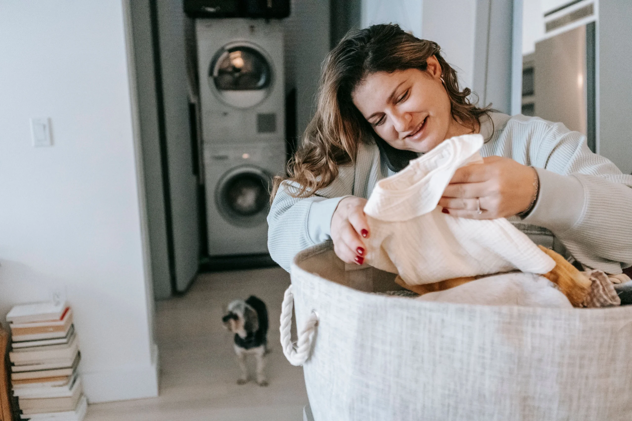 Woman folding fresh laundry from basket while phone rests on shoulder, with stacked washer and dryer visible in background — modern laundry services for busy households