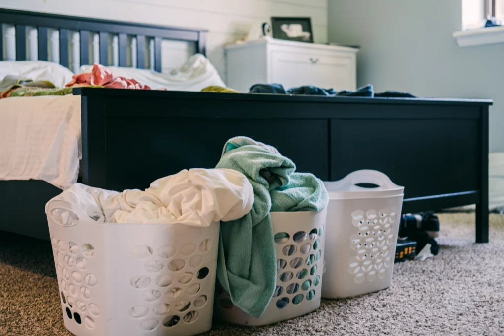 Three overflowing laundry baskets filled with sheets, towels, and clothes beside a bedroom bed — professional washing service laundry pickup for busy households